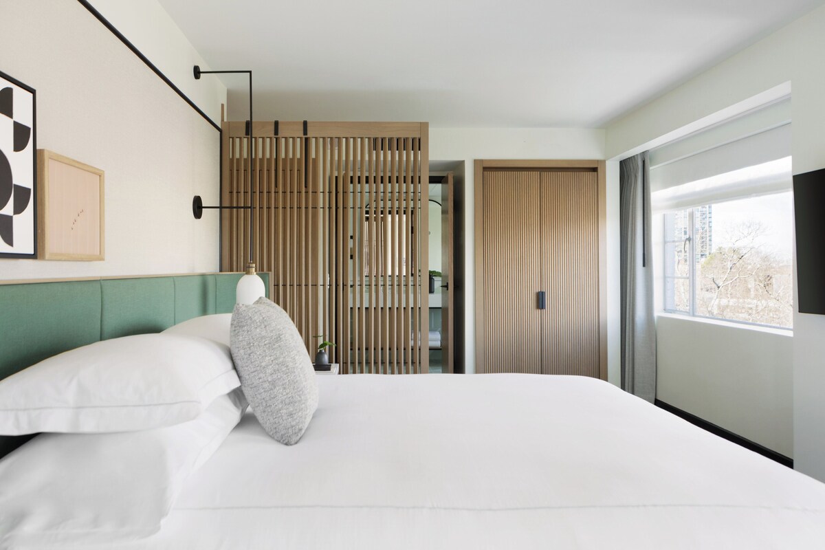 Een moderne hotelkamer met een groot bed, een groen hoofdeinde, een houten scheiding en een groot raam met uitzicht op gebouwen.