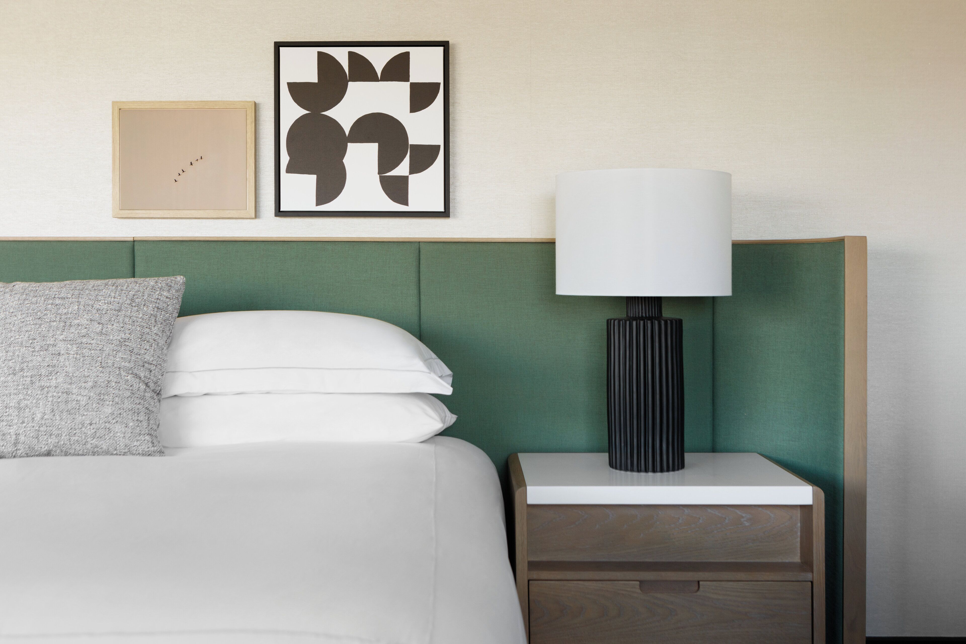 Un lit bien fait, avec du linge de lit blanc et un oreiller gris, une table de chevet avec une lampe, et deux œuvres d’art encadrées accrochées au mur.