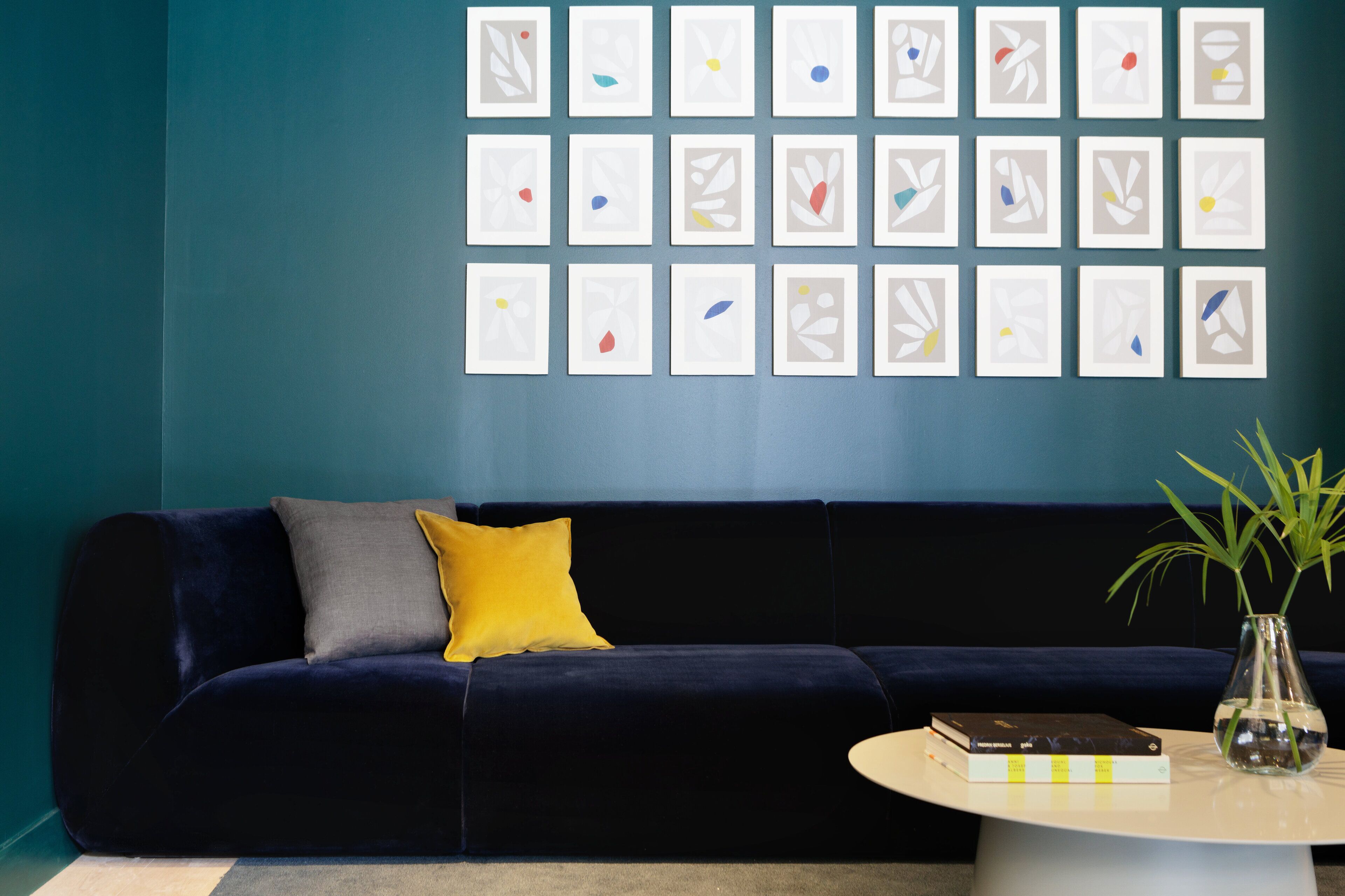 Een moderne woonkamer met een donkerblauwe bank, een geel kussen en een witte salontafel met boeken en een vaas met groene planten.