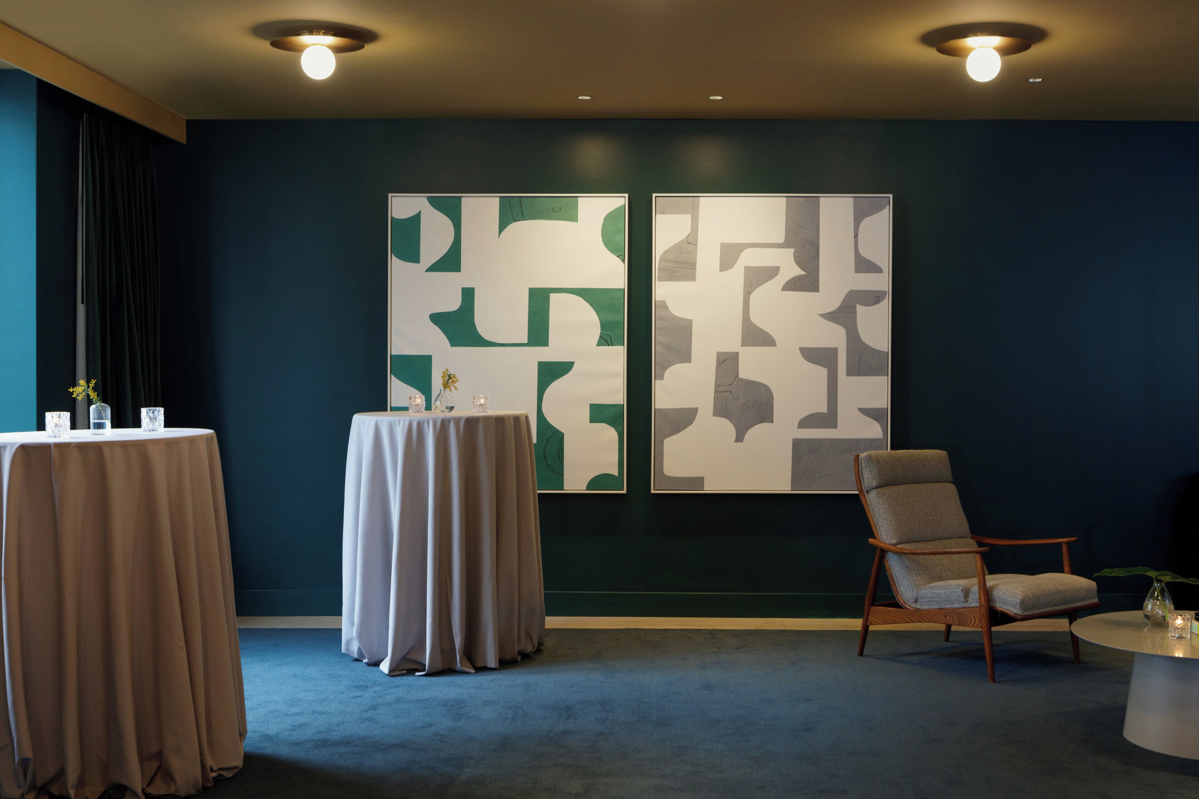 A room with a dark blue wall, two abstract art pieces, a round table with a chair, and a small table with a vase and glasses.