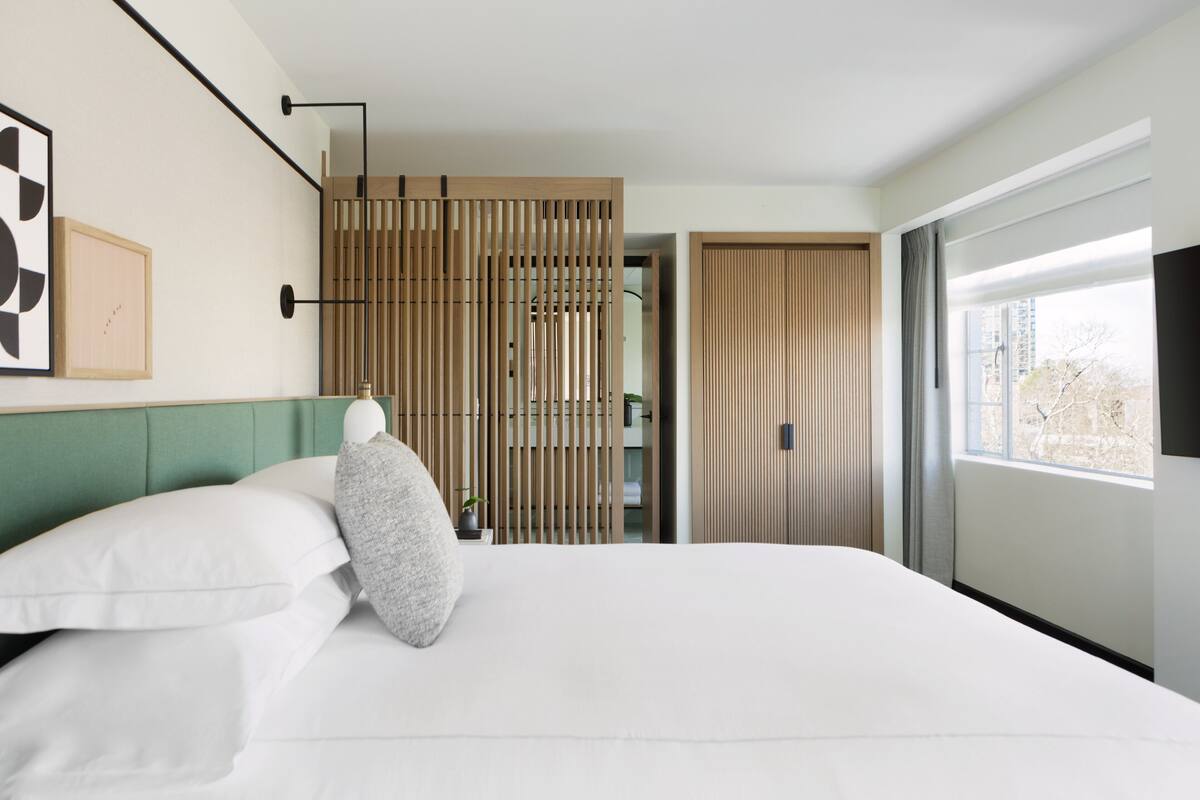 Moderni hotellihuone, jossa on suuri sänky, vihreä pääty, puinen sermi ja suuri ikkuna, josta avautuu näkymä rakennuksiin.