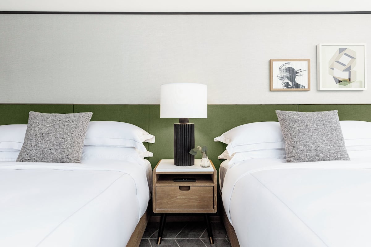 Hotellihuone, jossa on kaksi sänkyä, yöpöytä lampun kanssa ja taideteos seinällä.
