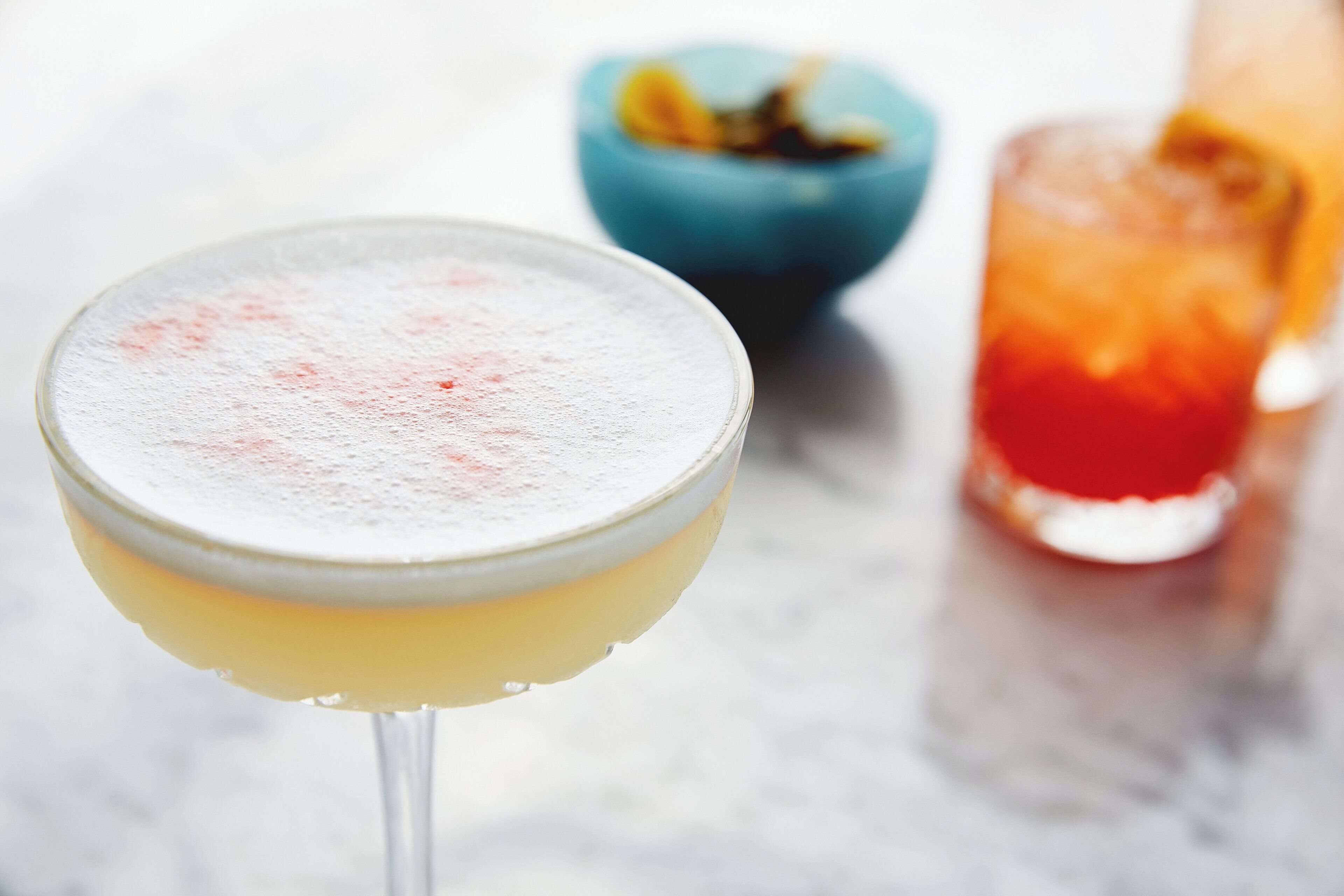 A cocktail glass with a frothy top, a bowl with a fruit garnish, and a glass with a red liquid.