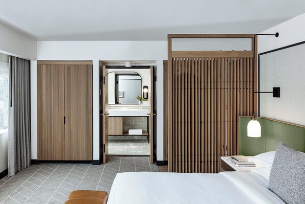 Een moderne hotelkamer met een bed, nachtkastje, spiegel en een badkamer met wastafel en handdoekhouder.