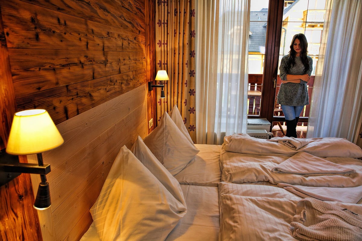 A hotel room with a wooden headboard, a bed with white linens, a lamp on the side table, and a view of the outside through the window.
