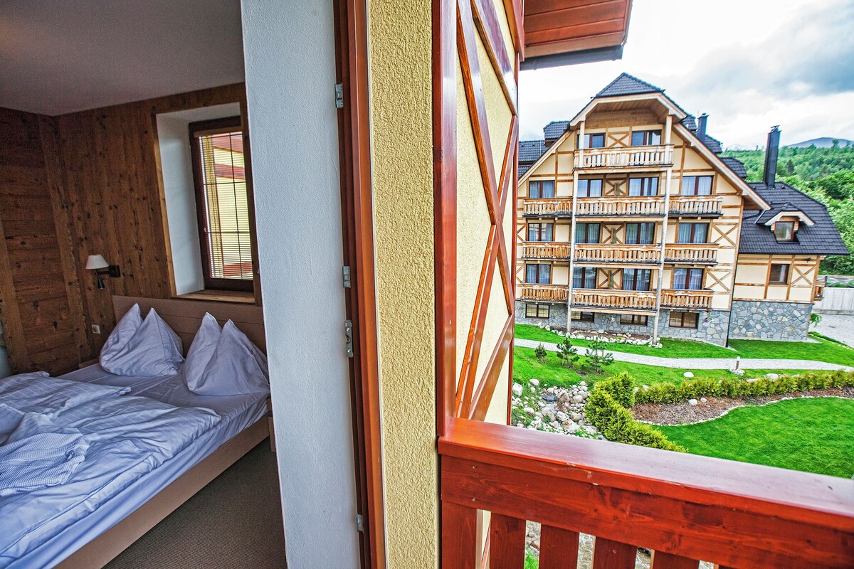A room with a bed, a window with blinds, and a view of a multi-story building with balconies.