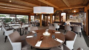 A restaurant with a central round table surrounded by chairs, a stone wall, and wooden beams on the ceiling.