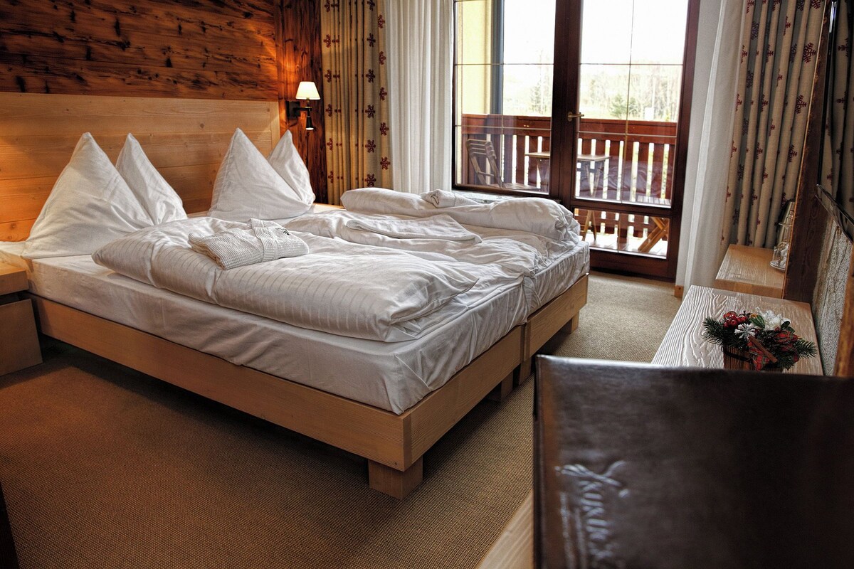 A hotel room with a wooden bed, a nightstand, a lamp, a window with a view of a balcony, and a small table with a decorative item.