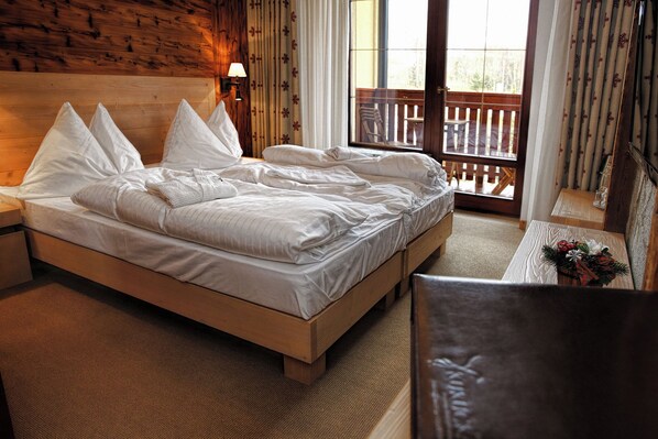 Habitación de hotel con cama de madera, mesita de noche, lámpara, ventana con vista a un balcón y una pequeña mesa con un objeto decorativo.