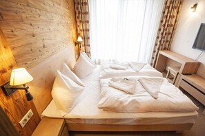 Habitación de hotel con cabecera de madera, lámpara de noche, cama con ropa blanca y televisor montado en la pared.