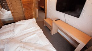 Un dormitorio con una cama, un banco de madera, un televisor montado en la pared y una puerta que da a otra habitación.