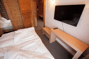 Un dormitorio con una cama, un banco de madera, un televisor montado en la pared y una puerta que da a otra habitación.