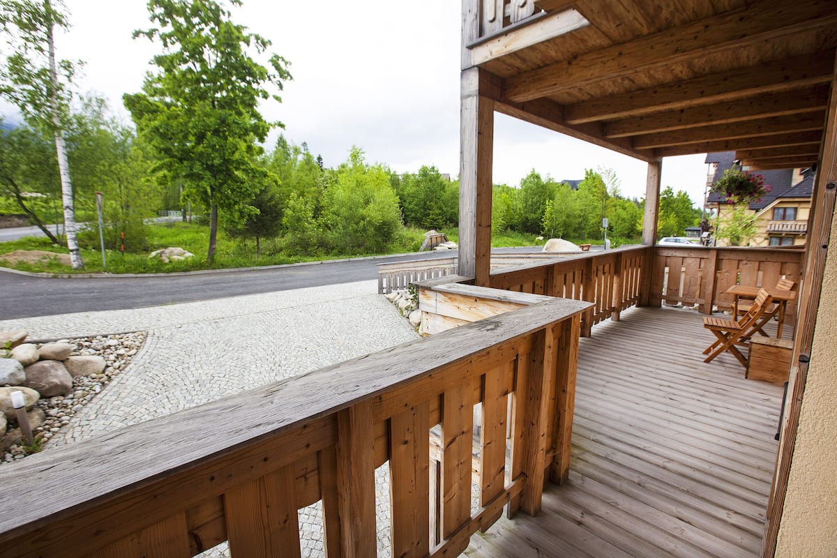 A wooden deck with a stone patio, outdoor seating, and a view of a road and greenery.