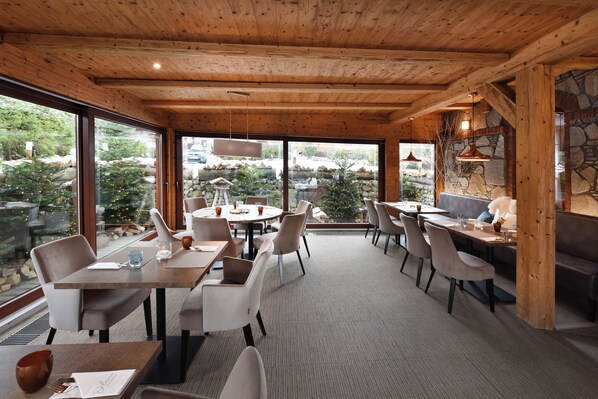 Un restaurante acogedor con techos de madera y grandes ventanales que ofrecen vistas al exterior. Las mesas están dispuestas con sillas y presentan manteles pequeños y velas.