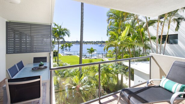 A balcony with a glass railing, a dining table with chairs, and a view of a body of water and palm trees. - Noosa Shores (Noosa Heads)
