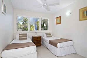A room with two single beds, a ceiling fan, a nightstand with a lamp, and a window with blinds.