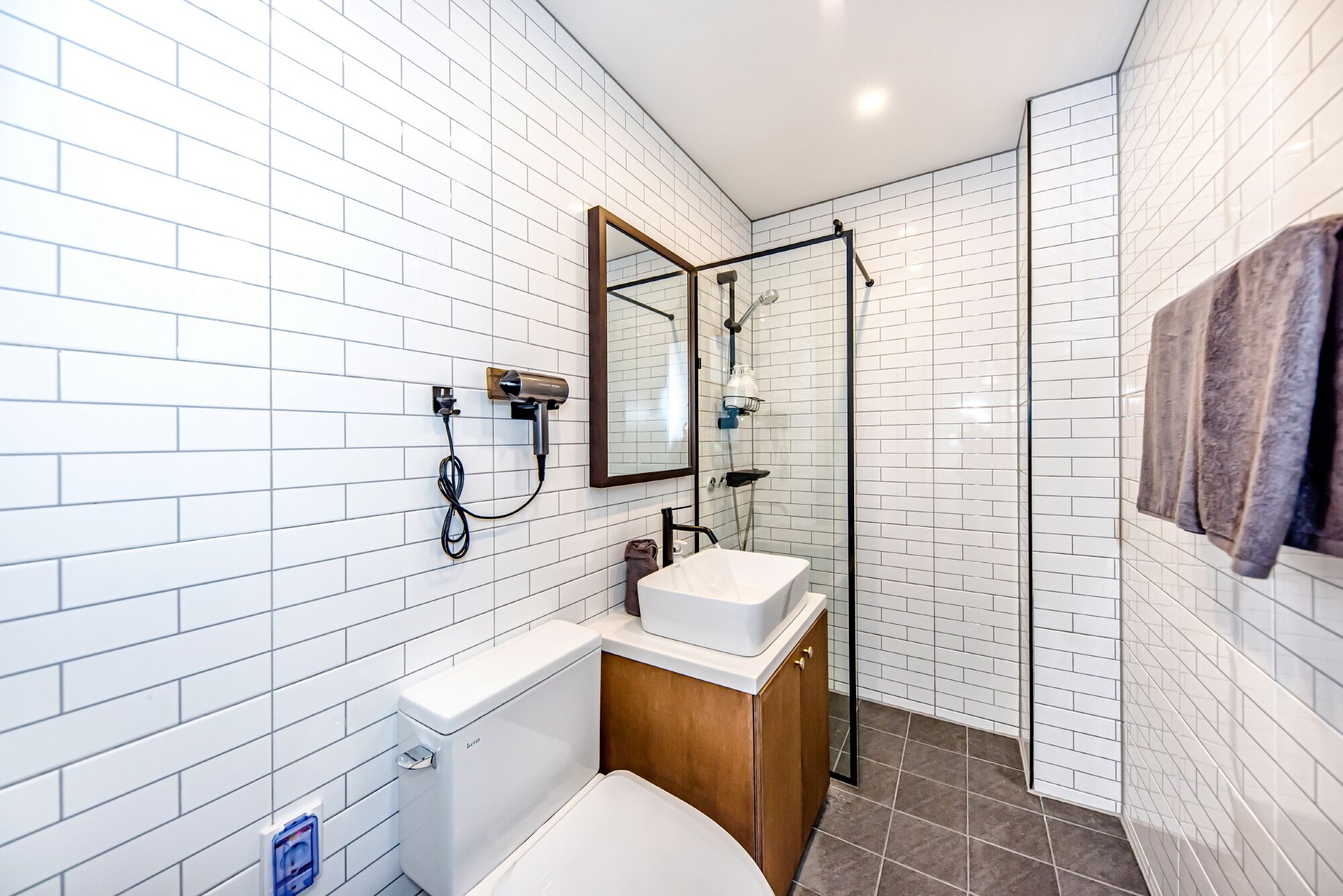 A modern bathroom with white subway tiles, a white toilet, a wooden vanity with a white sink, a glass shower enclosure, and a towel hanging on the wall.