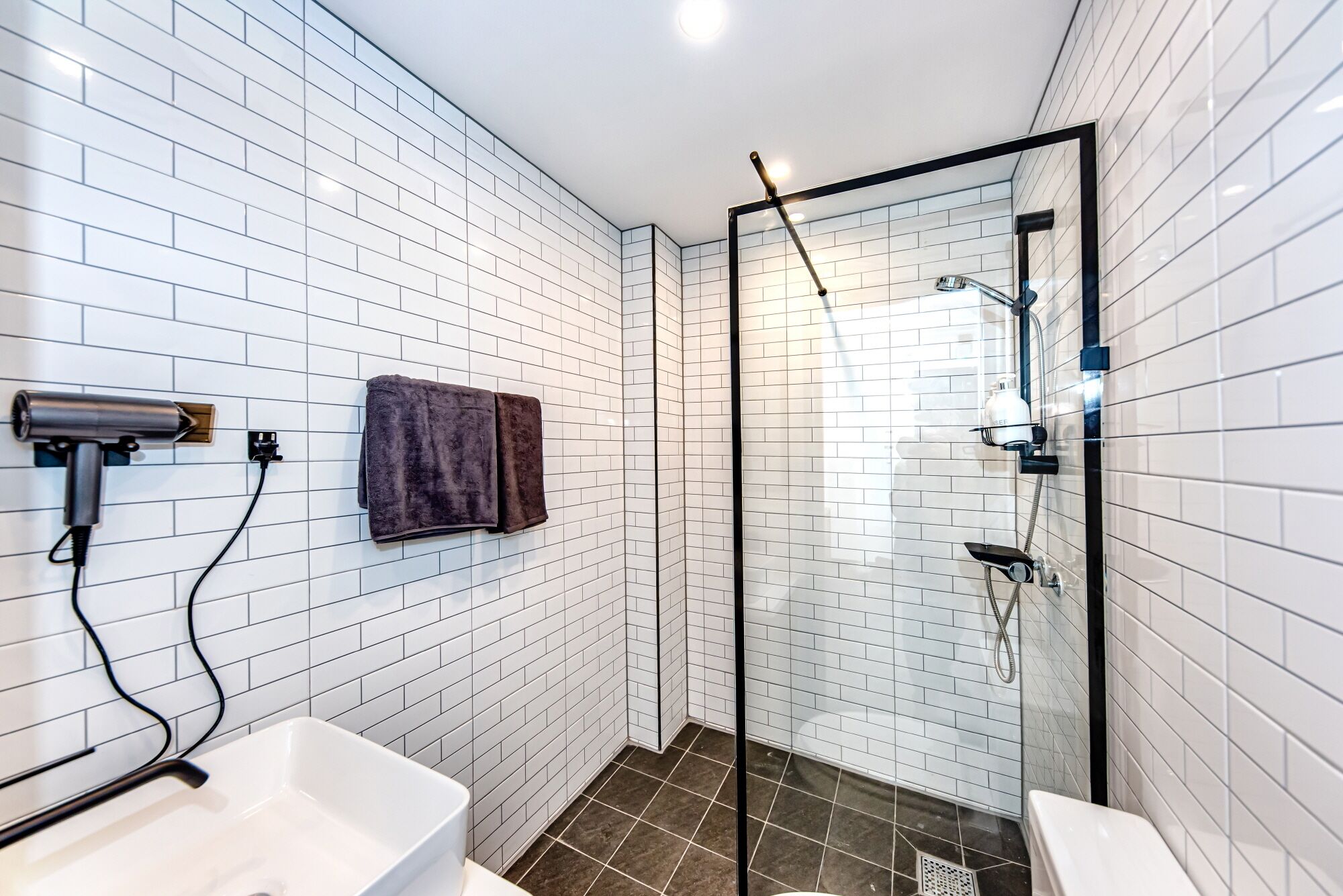 A modern bathroom with white subway tiles, a glass-enclosed shower, a white sink, and a dark towel hanging on the wall.