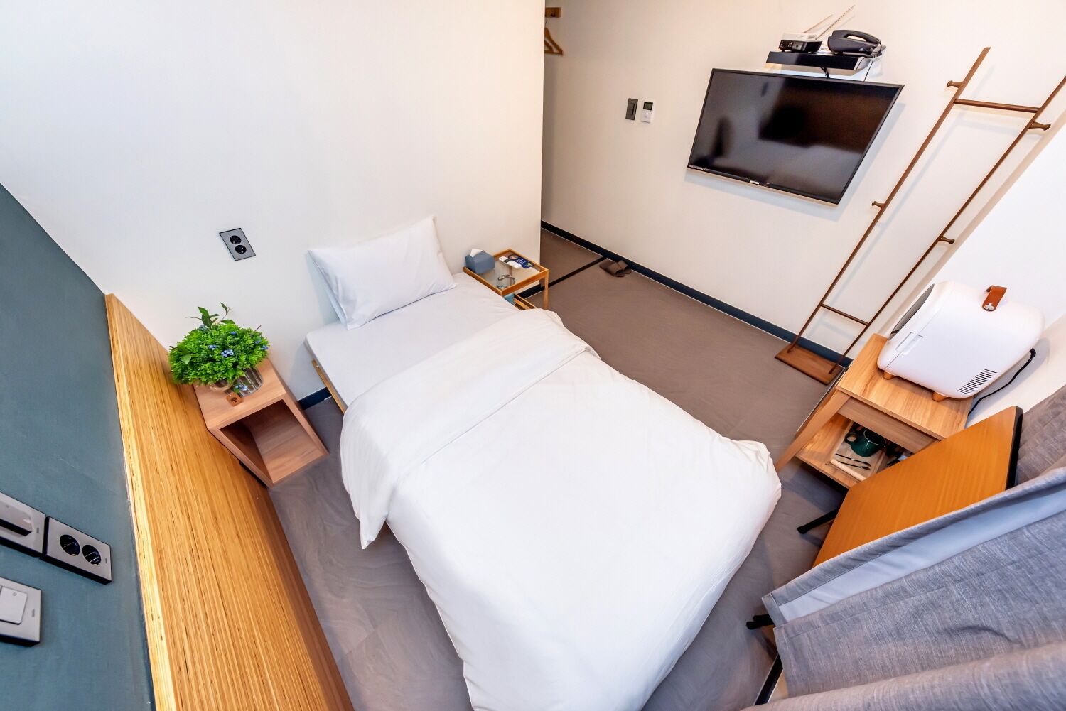 Une chambre d’hôtel moderne avec un lit simple, un téléviseur fixé au mur, une petite table de chevet, une lampe de chevet et un purificateur d’air fixé au mur.