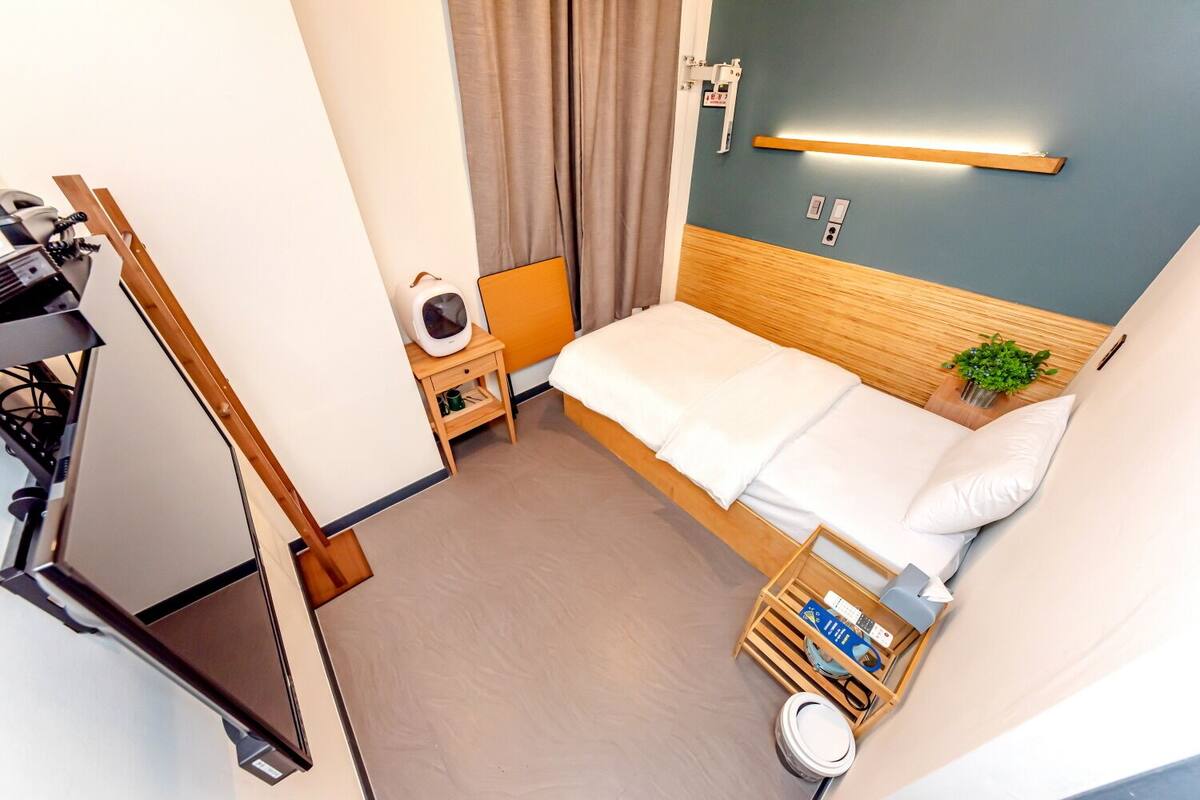 Een hotelkamer met een bed, een nachtkastje met een lamp, een kleine plant en een houten bijzettafeltje.