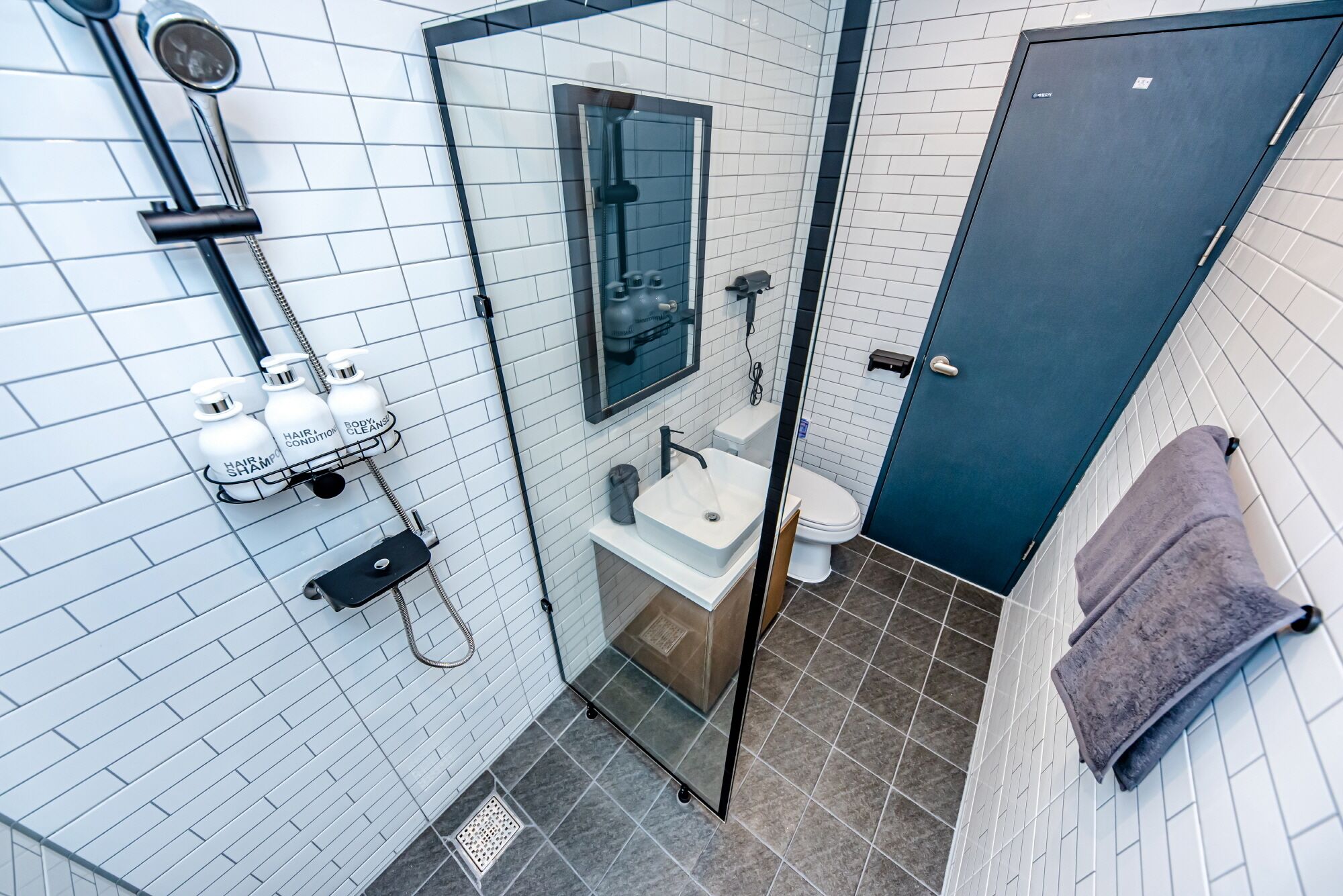 A modern bathroom with a shower, sink, and toilet. There are toiletries on a shelf, and a towel is hanging on the wall.