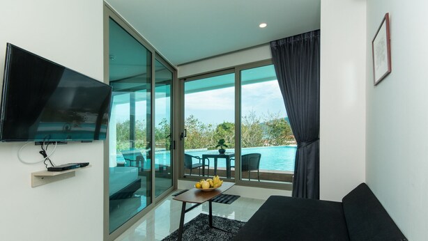 Uma sala de estar moderna com televisão, sofá, mesa de jantar com frutas e uma grande janela com vista para uma piscina e vegetação.