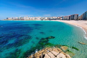 A coastal cityscape with a clear blue sea, sandy beach, and a row of modern buildings along the shore.