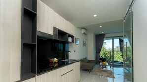 A modern kitchen with a black countertop, wooden cabinets, and a built-in oven. There is a dining area with a glass table and chairs, and a balcony with a view of greenery. - Luxury Sea View 1Bedroom Apartment (Kamala)