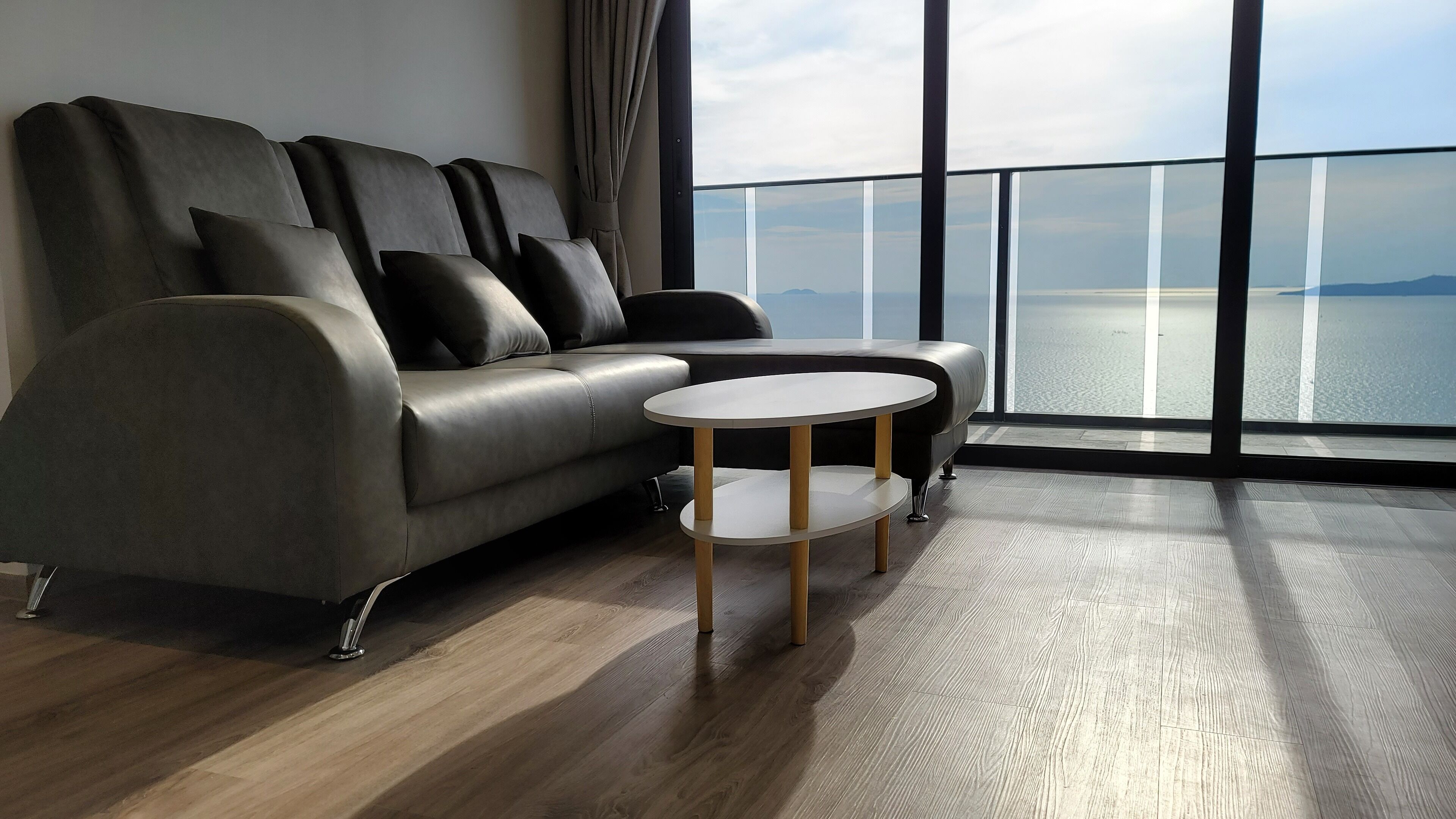 A modern living room with a grey sofa, a small round table, and a large window offering a view of the sea.
