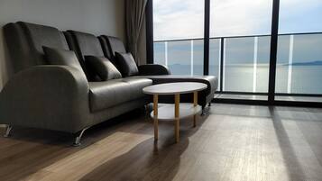 A modern living room with a grey sofa, a small round table, and a large window offering a view of the sea.