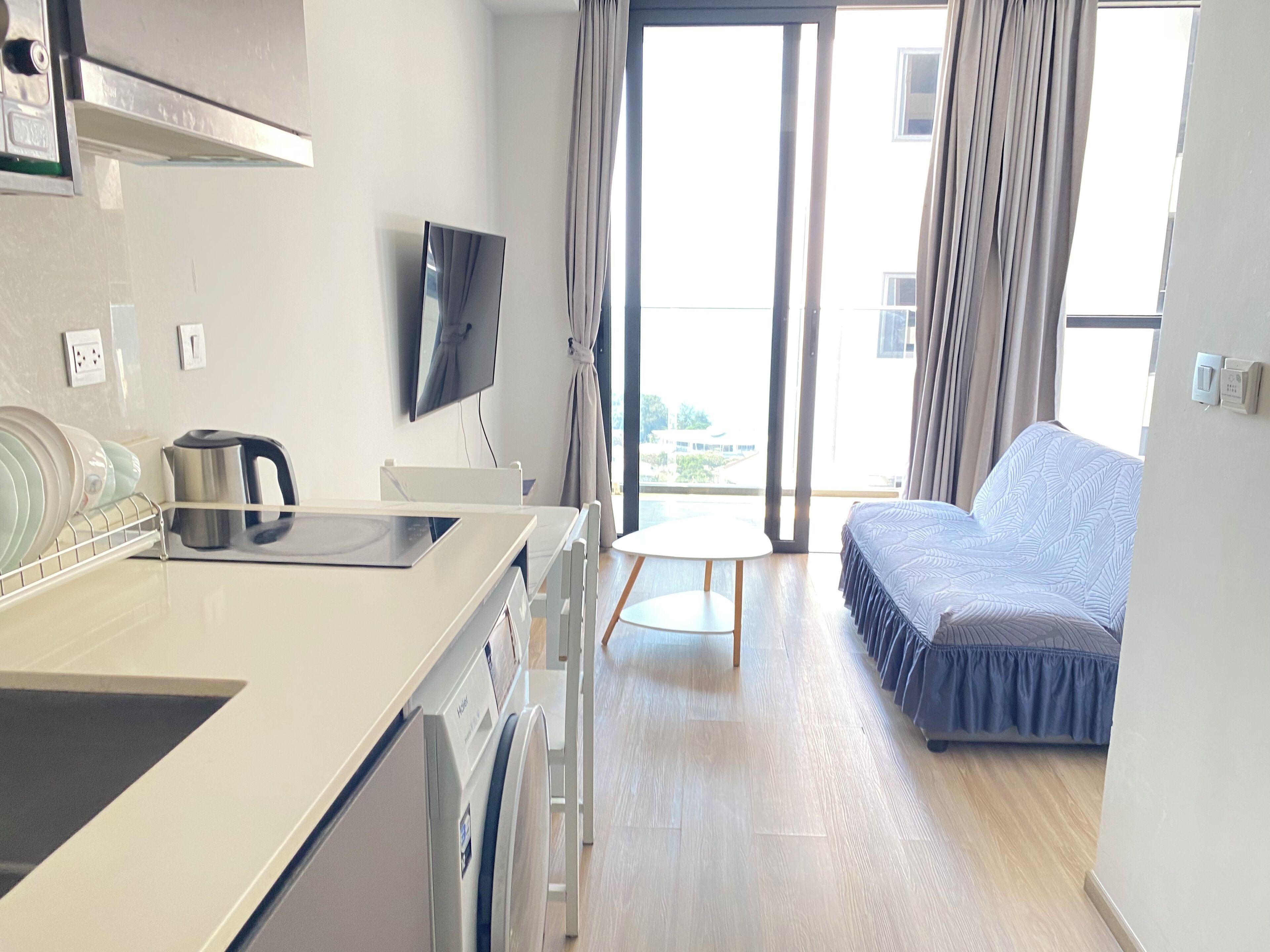A modern hotel room with a kitchenette, a flat-screen TV, a sofa, and a view of the outdoors.