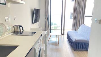A modern hotel room with a kitchenette, a flat-screen TV, a sofa, and a view of the outdoors.