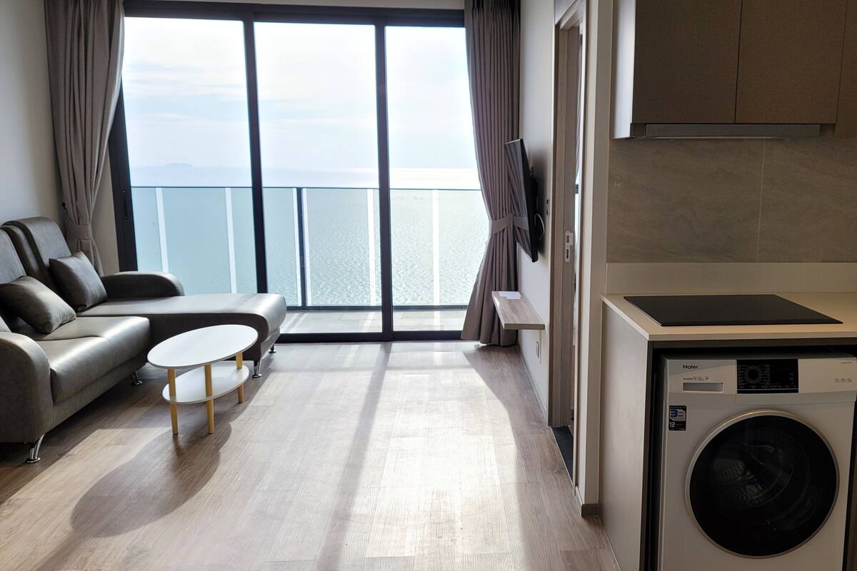 A modern living room with a sofa and coffee table, a kitchen with a washing machine, and a view of the sea through large windows.