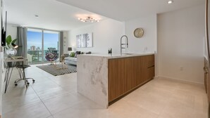 A modern kitchen with a marble island, wooden cabinets, and a large window offering a city view.