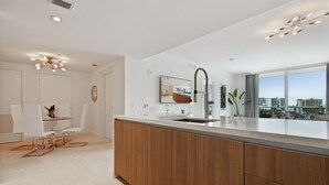 A modern kitchen with a central island, a dining area with white chairs, and a large window offering a city view.