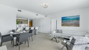 A modern living room with a dining area, white furniture, and a beach-themed painting on the wall. - Tiffany Ebony: 11th FL,Ocean-Canal views, Walk to Beach (Fort Lauderdale)