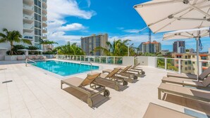 A rooftop pool area with lounge chairs and umbrellas, surrounded by high-rise buildings and palm trees. - Tiffany Ebony: 11th FL,Ocean-Canal views, Walk to Beach (Fort Lauderdale)