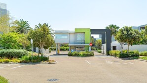 Um edifício moderno com fachada verde e cinza, área de estacionamento com via livre e vegetação exuberante ao redor.