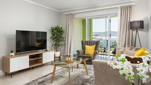 Uma sala de estar moderna com uma TV de tela plana, uma mesa de centro de vidro, um sofá com almofadas amarelas e uma janela grande com cortinas.