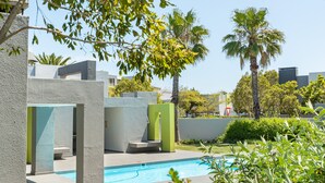 Uma área de piscina moderna ao ar livre, com estruturas geométricas em concreto, palmeiras e um céu azul claro.