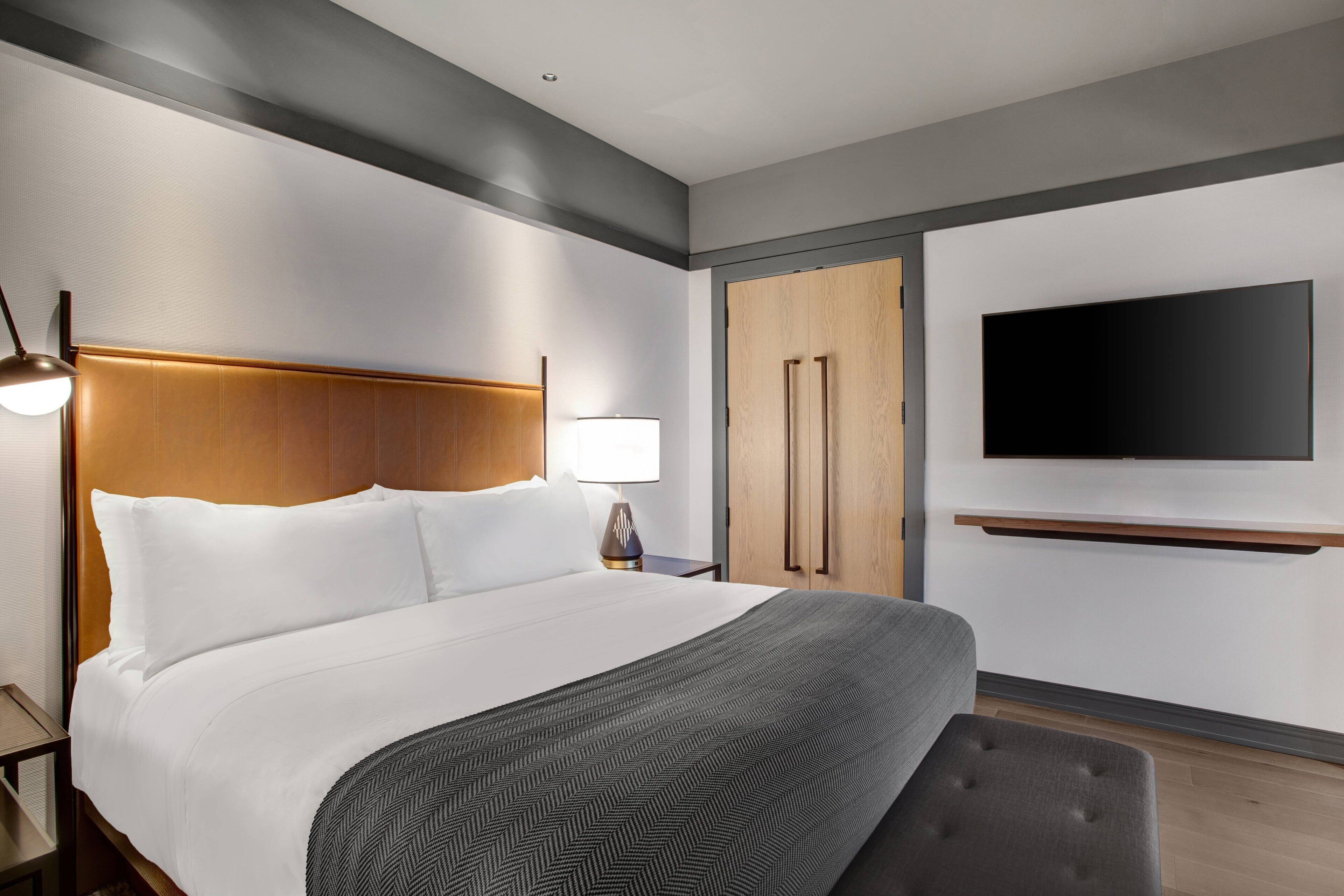 A modern hotel room with a large bed, a flat-screen TV mounted on the wall, a wooden wardrobe, and a bench.