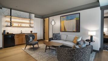 Una sala moderna con sofá, sillón, mesa de centro y una zona de cocina con estanterías y refrigerador.