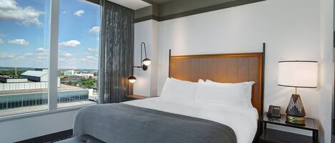 Hotelkamer met een groot bed, een bankje, een nachtkastje met een lamp en uitzicht op de stad door grote ramen.