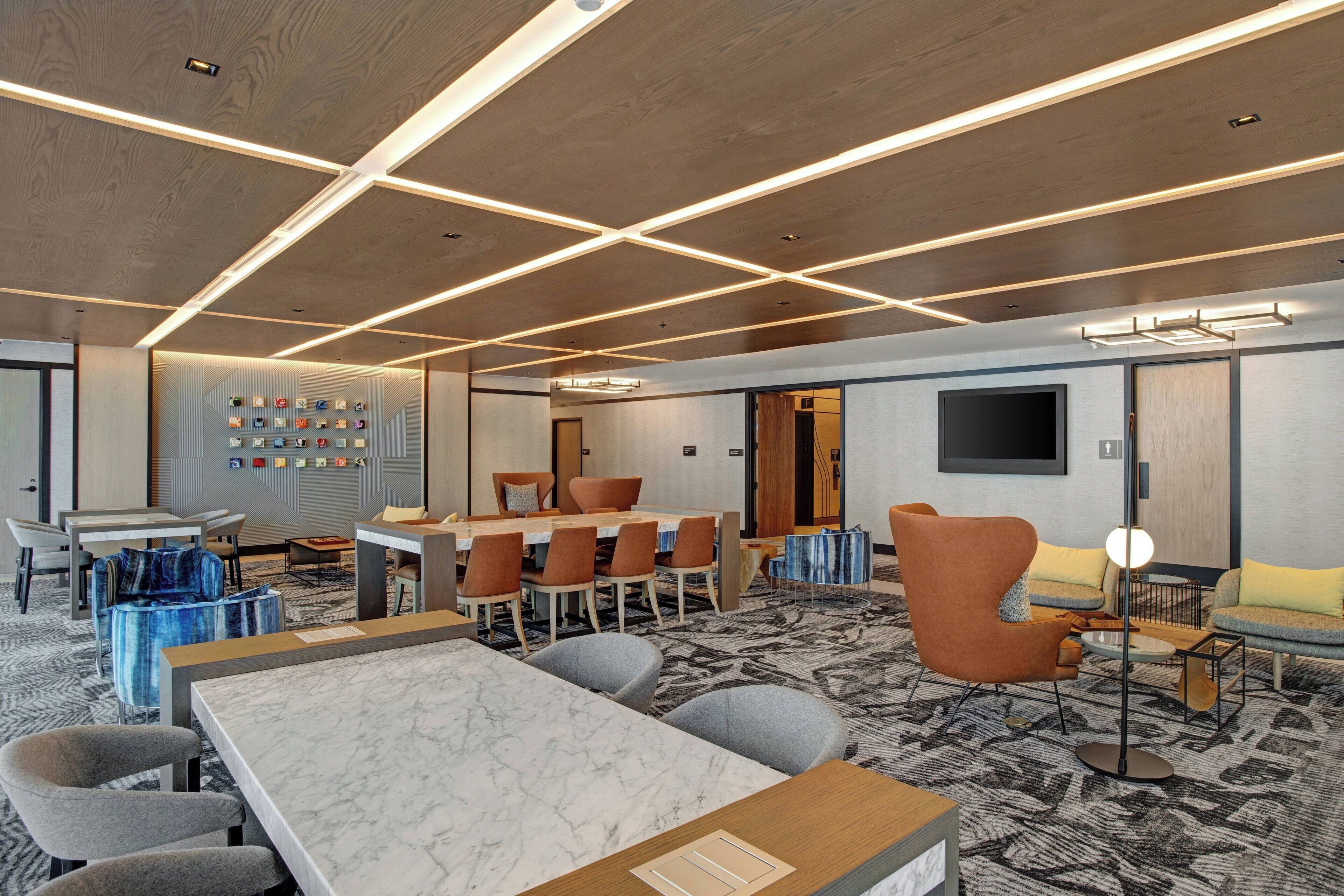 A modern hotel room with a marble-topped dining table, comfortable seating, a flat-screen TV, and a wall-mounted clock.