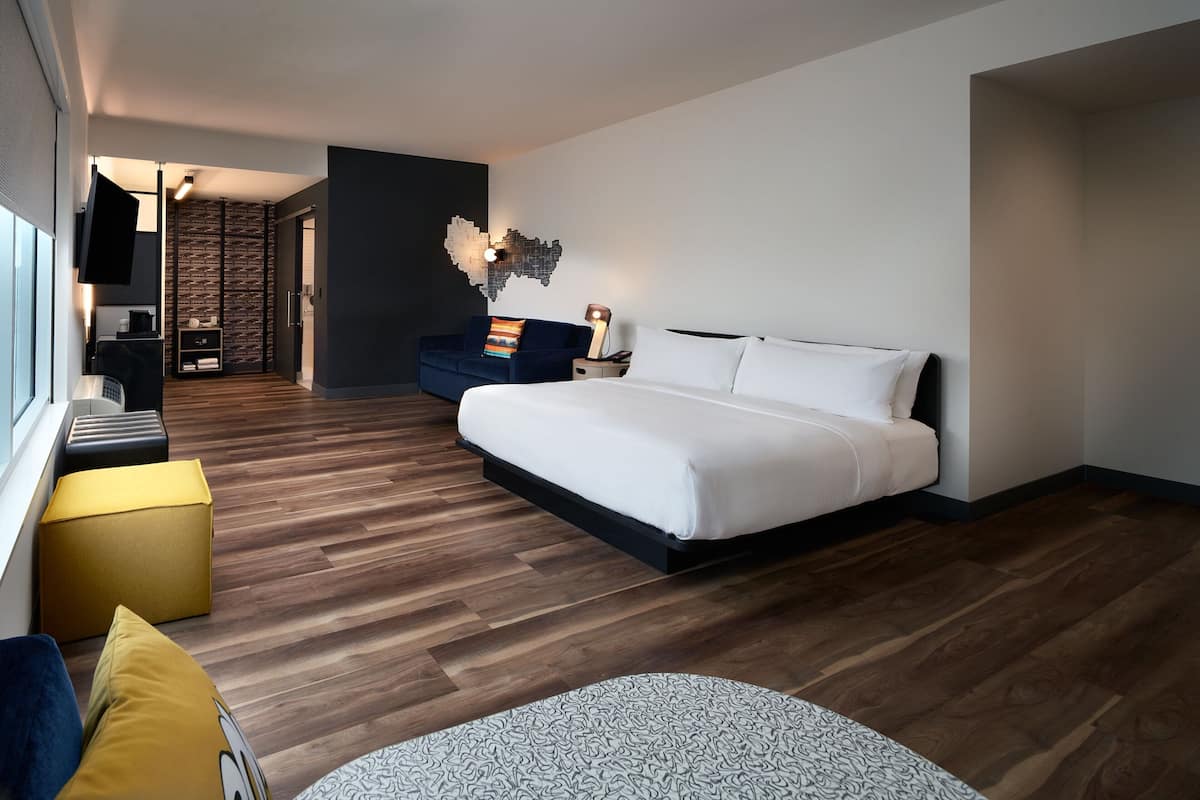 Moderni hotellihuone, jossa on suuri sänky, sohva, työpöytä tuolilla ja seinälle asennettu televisio.