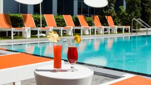 Un bar junto a la piscina con dos bebidas, sillones reclinables anaranjados y un edificio al fondo.