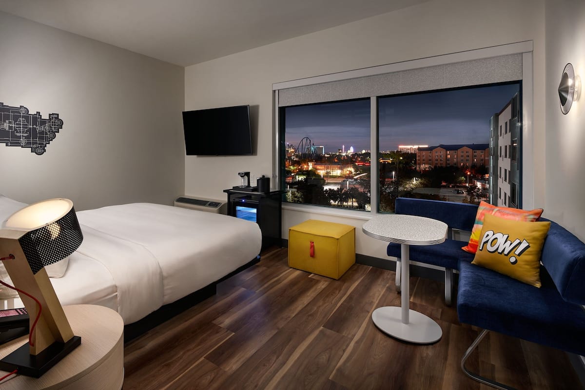 Moderni hotellihuone, jossa on sänky, työpöytä, tuoli, televisio ja suuri ikkuna, josta avautuu näkymä kaupunkiin.
