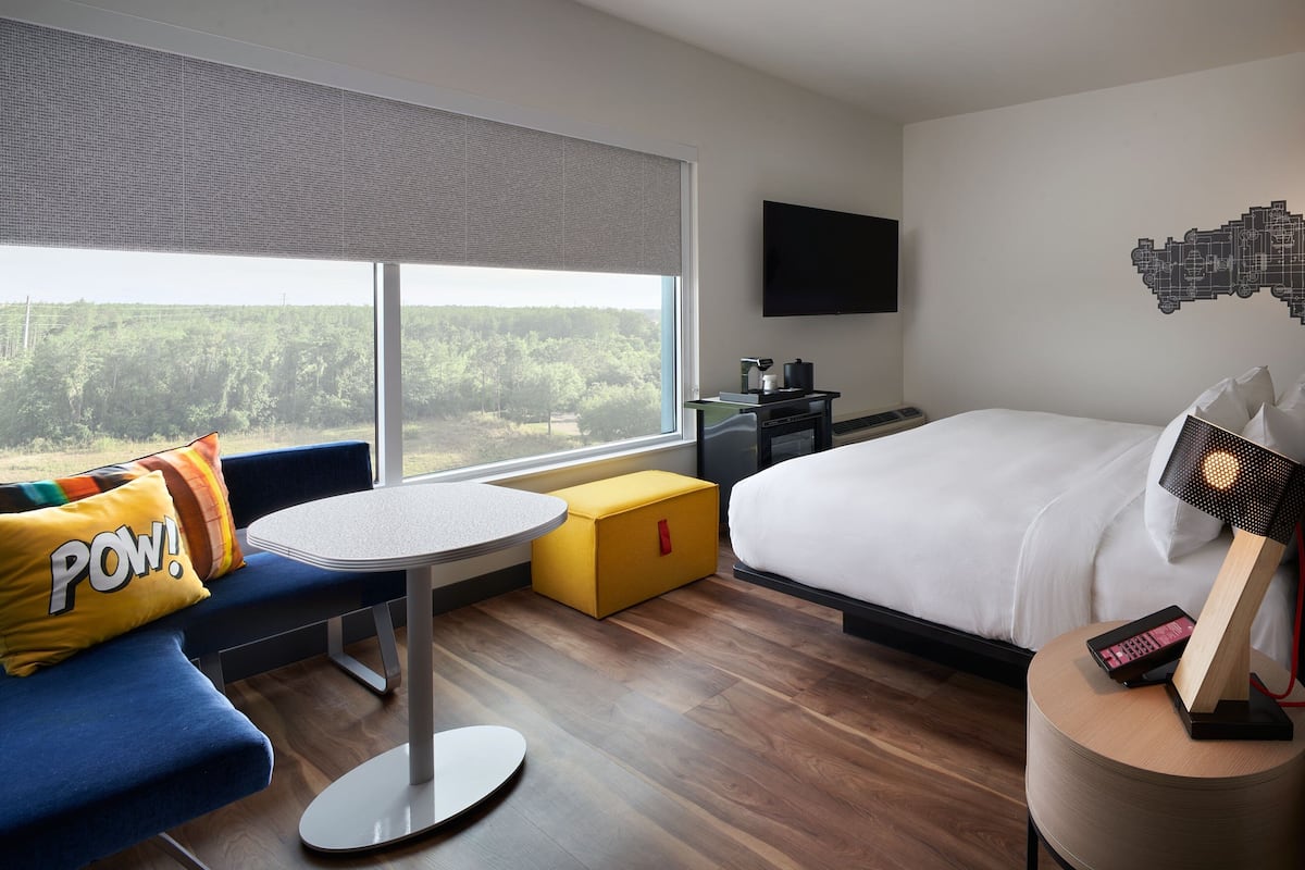 Hotellihuone, jossa on suuri ikkuna, sänky, työpöytä, tuoli ja sohva, jonka päällä on koristeellinen tyyny, jossa on teksti »POW!».