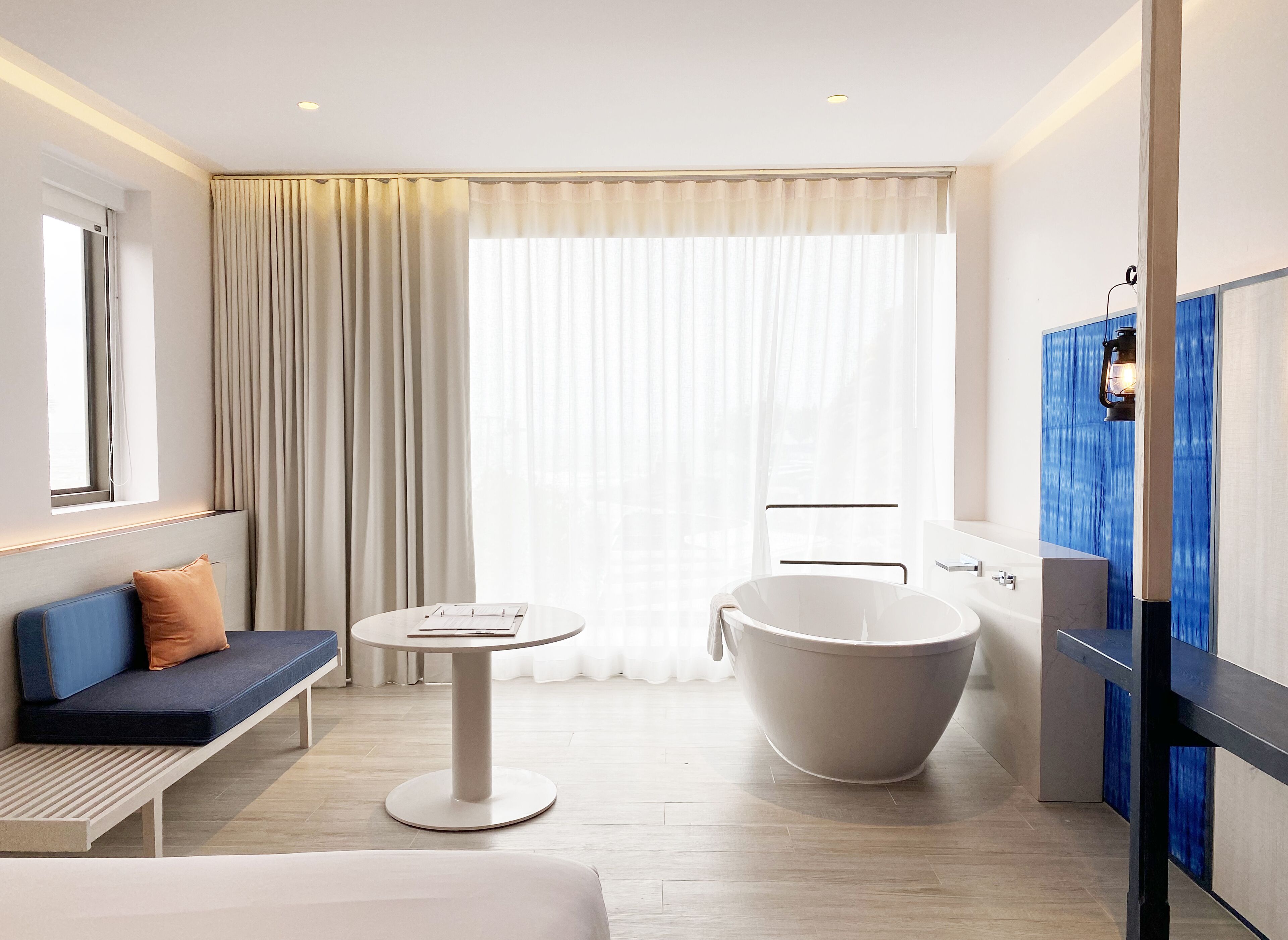 A modern hotel bathroom with a freestanding bathtub, a round table, and a bench with cushions.