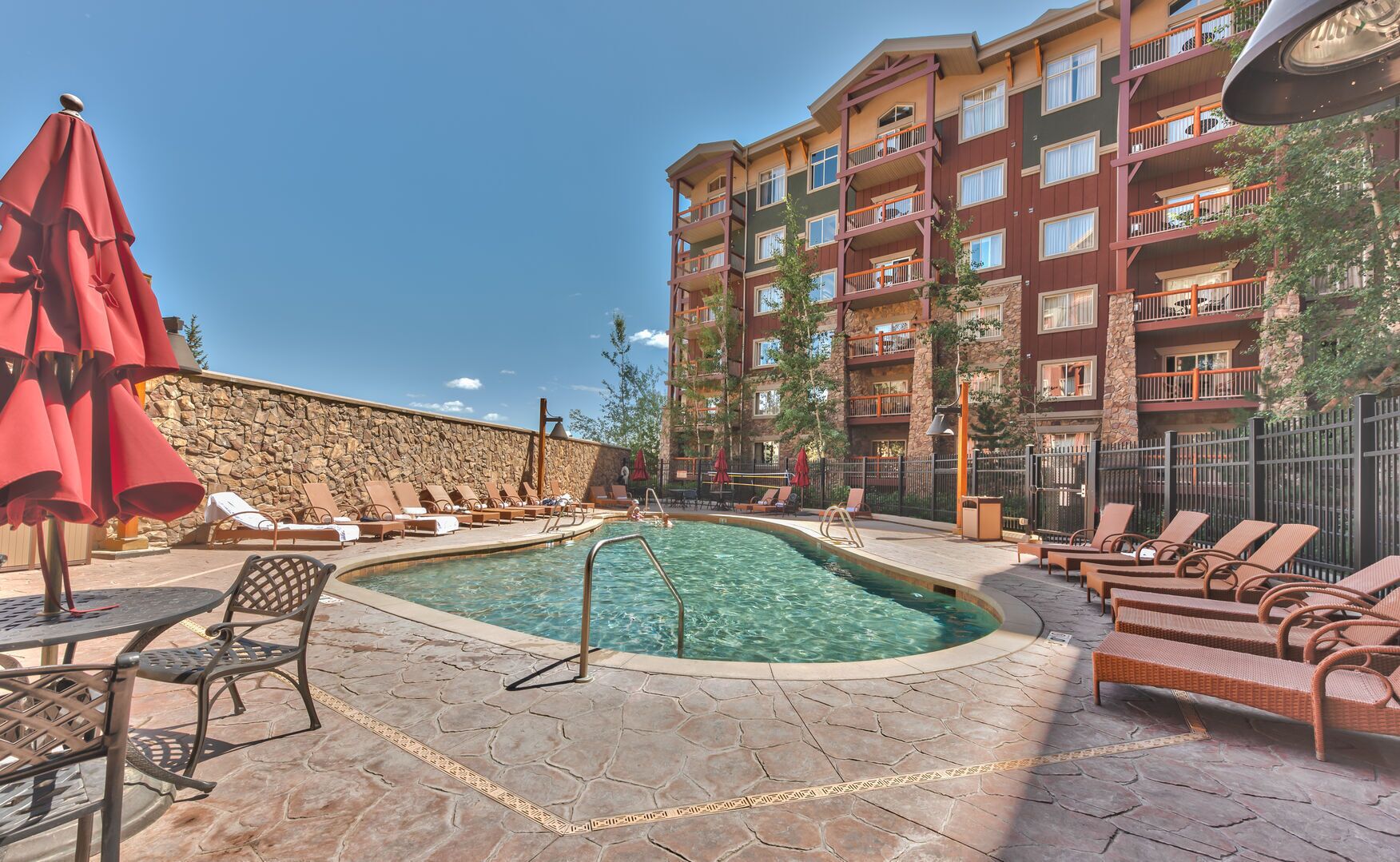 A pool area with lounge chairs and umbrellas, a stone wall, and a multi-story building with balconies.